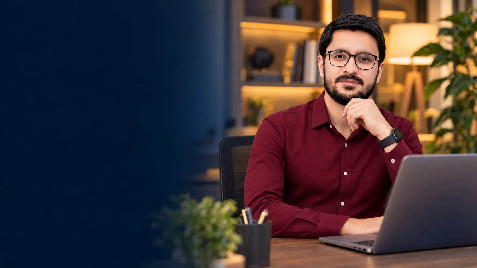 Shahzad Khan wearing glasses and a burgundy shirt, working at a laptop in a warm modern office.